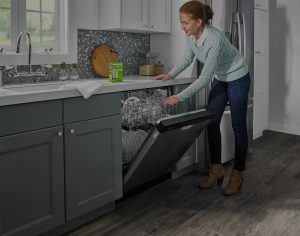 A box of affresh® dishwasher cleaner tablets sits on the counter while a woman inspects the clean, residue-free glasses she has taken from her dishwasher's top rack.