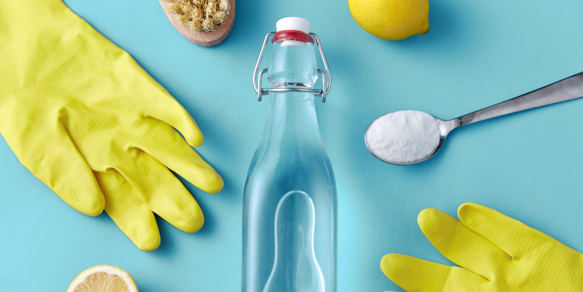 Horizontal photo on a soft blue background featuring a spoonful of baking soda, yellow cleaning gloves, a scrub brush partially visible, a lemon, and a central bottle.