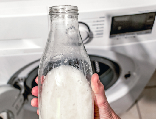 A glass bottle filled with vinegar being held in front of a washing machine for use in sanitization.