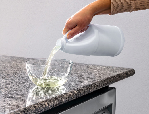 Pouring bleach into a clear bowl on a kitchen counter above the dishwasher, illustrating the process of preparing to sanitize a dishwasher with bleach.