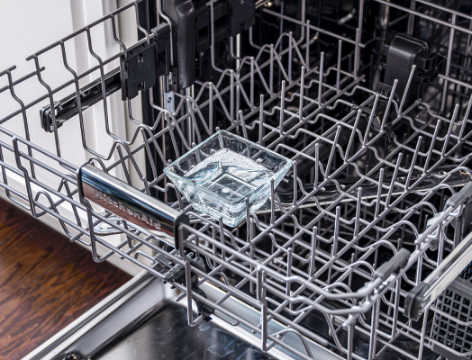 Open dishwasher with a dishwasher-safe container filled with vinegar on the top rack, illustrating the process of sanitizing a dishwasher using vinegar.